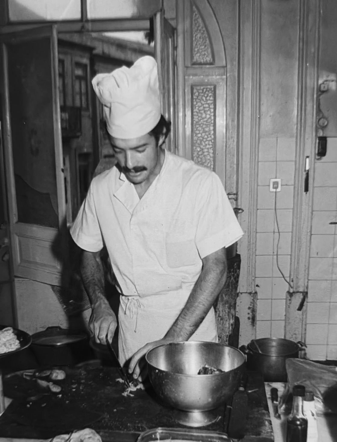 José Magalhães, jovem, de toque e jaleca, a trabalhar numa cozinha de azulejo